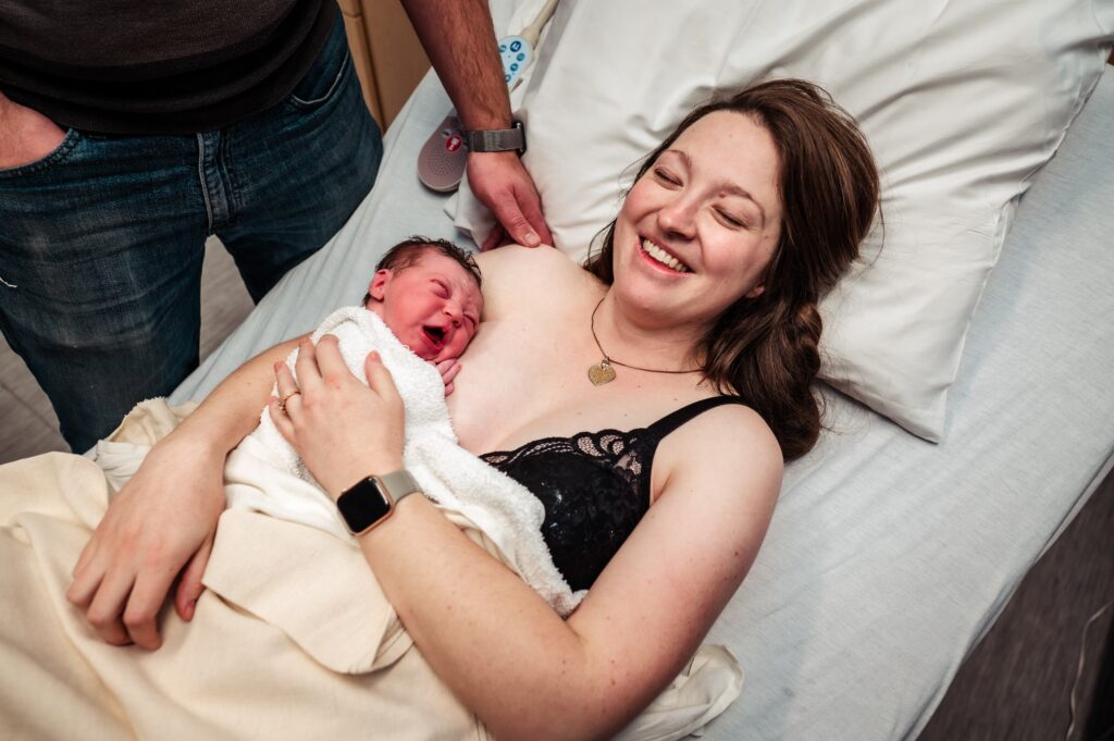 Kaitlyn on a hospital bed, sitting in the contagious joy minutes after Emma's birth, her husband's hand visible on her shoulder, Emma (the babe) crying on her breast.