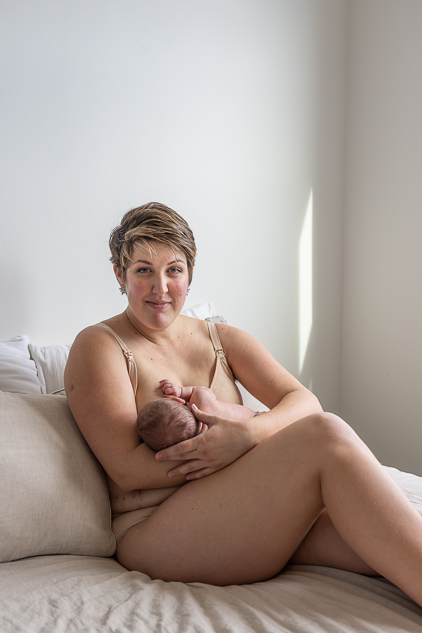 An intimate postpartum breastfeeding portrait; mom is sitting on a soft linen bed, wearing nude underwear and looks directly at the camera as her baby feeds at her breast. She is confident and brimming with self-love.